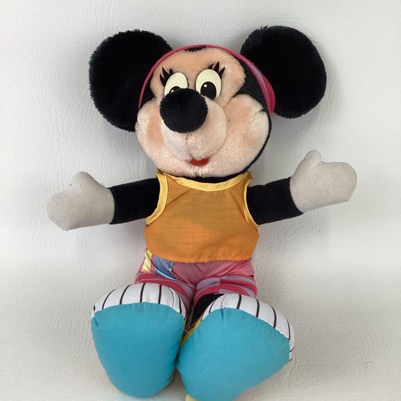 May include: A plush toy of a cartoon mouse with large black ears, a peach-colored face, and long eyelashes. The character wears a yellow sleeveless top, pink and purple striped pants, and large bright blue shoes with white pinstriped accents.