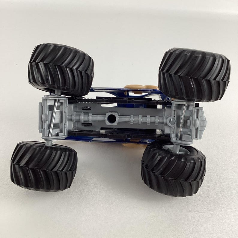May include: A blue and gray toy monster truck with four large black tires. The underside of the truck is visible, showing the chassis and suspension details. The tires have a deep tread pattern.