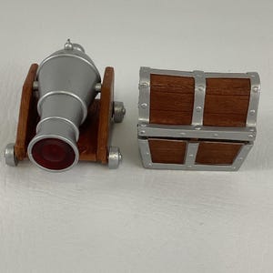 May include: A miniature cannon and treasure chest set. The cannon is silver with a red opening and sits on a brown wooden base. The treasure chest is brown with silver metal accents. The set is designed for decorative purposes.
