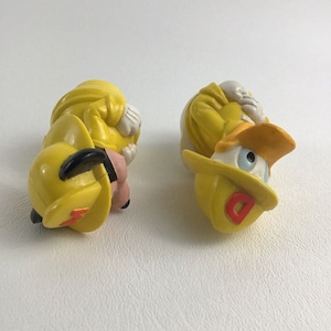 May include: Two yellow plastic figurines of cartoon characters wearing firefighter hats. One figurine is of a dog with a red '1' on its hat, and the other is of a duck with a red 'D' on its hat.