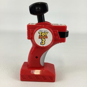 May include: A red and black Fisher-Price Toy Story 3 remote control toy. The toy has a handle, a black joystick, and a circular emblem with the Toy Story 3 logo. The base of the toy is red and rectangular.