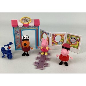 Peut inclure: Un ensemble de jouets Peppa Pig avec trois figurines de personnages, un tricycle bleu, un magasin de jeu et un présentoir pliable. Les figurines comprennent un cochon en robe rouge, un cochon en robe jaune et un personnage avec un casque. Un jeu de marelle est également inclus.