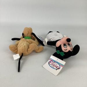 May include: Two plush toys featuring a tan dog with a green collar and a black, white, and pink mouse character wearing a green leaf crown. The mouse toy has a white tag attached that reads The Disney Store in blue and red lettering.