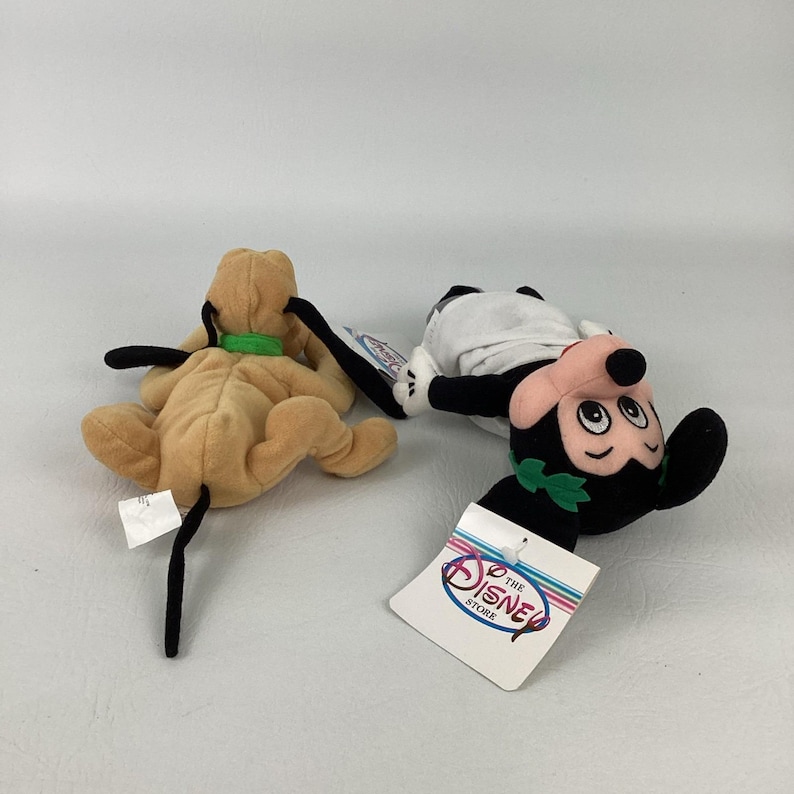 May include: Two plush toys featuring a tan dog with a green collar and a black, white, and pink mouse character wearing a green leaf crown. The mouse toy has a white tag attached that reads The Disney Store in blue and red lettering.