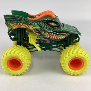 Pu&ograve; includere: Un giocattolo di monster truck verde e arancione con un design a testa di drago. Il camion ha grandi pneumatici gialli con centri arancioni e una carrozzeria verde dettagliata. La testa di drago presenta corna arancioni e un'espressione minacciosa.