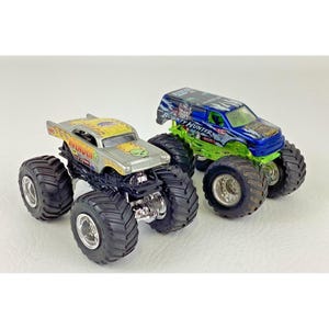 K&ouml;nnte beinhalten: Zwei Spielzeug-Monstertrucks. Einer ist silber und gelb mit dem Wort "Avenger" an der Seite. Der andere ist blau und gr&uuml;n mit den Worten "Bounty Hunter". Beide haben gro&szlig;e schwarze Reifen.