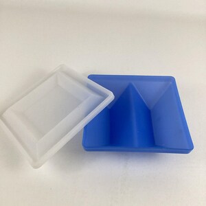 May include: A white square lid and a blue square tray with a pyramid-shaped indentation in the center.