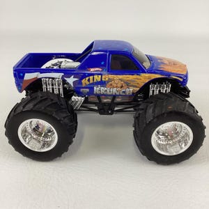 Op de afbeelding: Een blauwe Hot Wheels monstertruck speelgoed met grote zwarte banden en chromen velgen. De truck heeft de tekst "KING KRUNCH" en een afbeelding van een gespierde man. De truck heeft een blauwe cabine met sterren en vlammen.