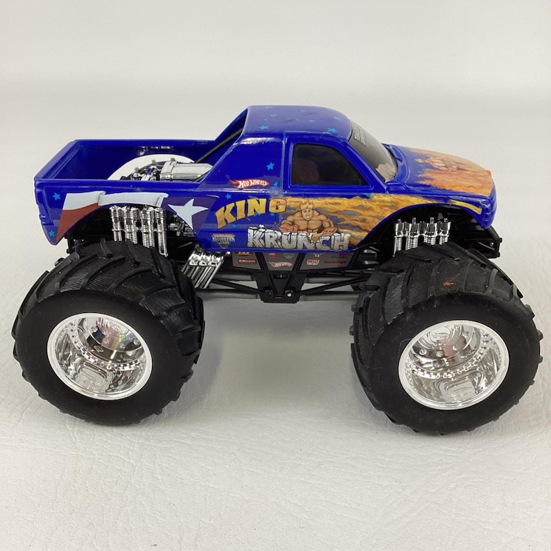Op de afbeelding: Een blauwe Hot Wheels monstertruck speelgoed met grote zwarte banden en chromen velgen. De truck heeft de tekst "KING KRUNCH" en een afbeelding van een gespierde man. De truck heeft een blauwe cabine met sterren en vlammen.