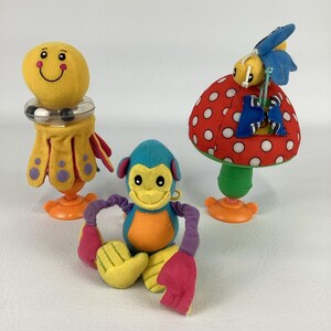 May include: Three colorful plush baby toys. One is a yellow smiling face with a silver rattle and yellow skirt. Another is a blue and orange monkey with purple arms and red hands. The last is a red mushroom with white spots.