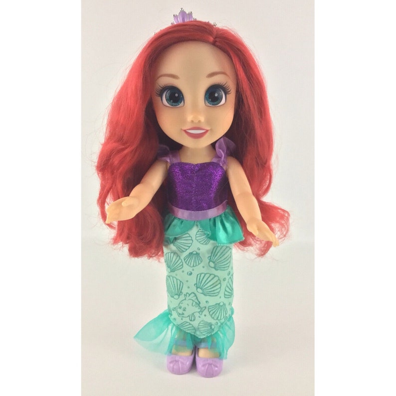 May include: A red-haired doll wearing a purple and green mermaid costume. The doll has a crown on her head and is smiling.