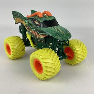 Pu&ograve; includere: Un giocattolo di monster truck verde e arancione con un design a forma di drago. Il camion ha grandi pneumatici gialli con cerchi arancioni. La carrozzeria del camion &egrave; verde con un volto e corna di drago dettagliati.