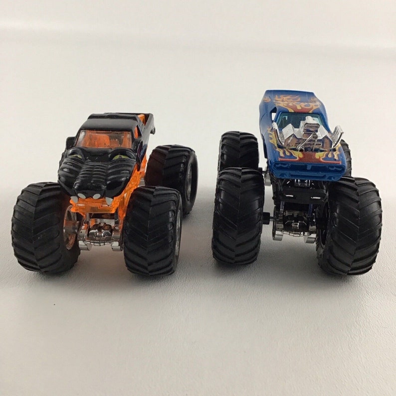 May include: Two toy monster trucks, one black with orange accents and a panther face design, and one blue with orange flames and a white interior. Both trucks have large black wheels.
