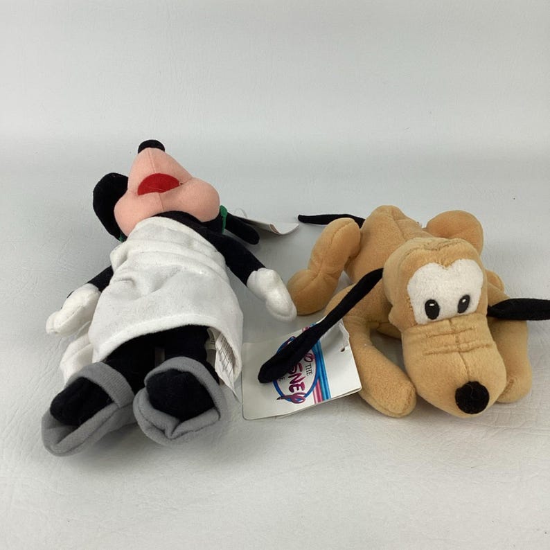May include: Two plush toys featuring a character with black ears, a pink face, and a white apron, alongside a tan dog character with long black ears and a black nose. Both toys have attached white tags with the Disney logo printed in blue and red.