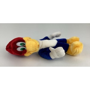 Op de afbeelding: Een pluche speelgoed van Woody Woodpecker, een stripfiguur. Het speelgoed heeft een rode kop, een gele snavel en groene ogen. Het draagt een blauw lichaam met een witte borst en witte handschoenen. De voeten zijn geel.