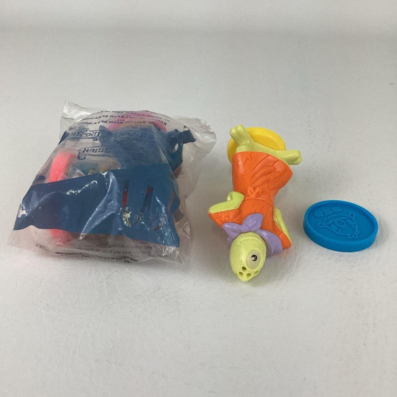 May include: A collection of children's toys, including a sealed plastic bag with a blue and pink toy inside, a yellow and orange figure with a purple bow, and a blue circular token with a cartoon character.