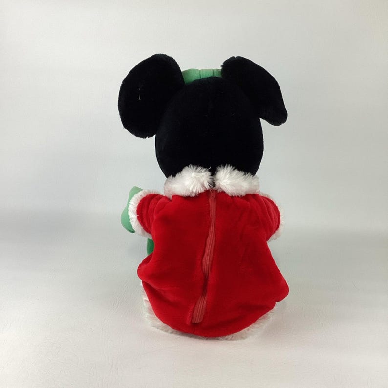 Puede incluir: Un peluche de un personaje negro con grandes orejas redondas, que lleva un abrigo de textura aterciopelada roja con ribete de piel sint&eacute;tica blanca en el cuello y el dobladillo. El abrigo tiene una cremallera vertical en la espalda y un objeto verde.