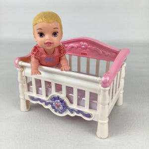May include: A plastic toy crib with a white frame, pink headboard, and purple floral accent, containing a small doll with blonde hair and a pink top. The Fisher-Price logo is visible on the headboard of the crib, which features a light purple mattress.
