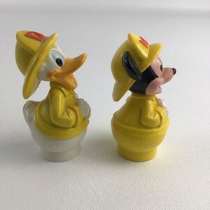 May include: Two yellow plastic figurines of Mickey Mouse and Donald Duck dressed as firefighters. Mickey is wearing a yellow hat with a red 'M' and Donald is wearing a yellow hat with a red 'D'.