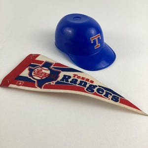 Op de afbeelding: Een blauw en wit vaandel met de woorden "Texas Rangers" en een honkbal met het woord "Rangers" erop. Een blauwe plastic honkbalpet met een rode "T" erop.