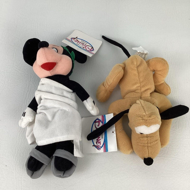 May include: Two plush toys featuring a character wearing a white toga, green laurel wreath, and grey sandals, alongside a tan dog character with black ears and a black nose. Both plush figures have attached Disney Store paper tags.