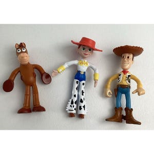 May include: Three plastic Toy Story character figurines: a brown horse, a cowgirl with a red hat and spotted pants, and a cowboy with a brown hat and vest. The figures are standing on a white surface.