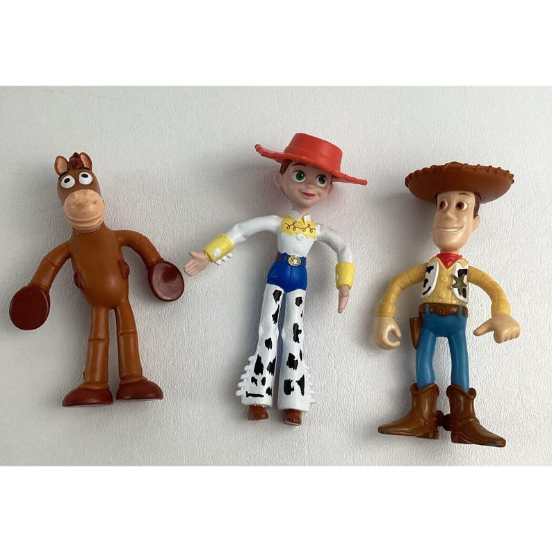 May include: Three plastic Toy Story character figurines: a brown horse, a cowgirl with a red hat and spotted pants, and a cowboy with a brown hat and vest. The figures are standing on a white surface.