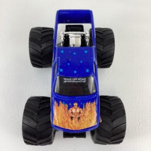 Op de afbeelding: Een blauw monstertruckspeelgoed met grote zwarte banden. De truck heeft een vlammenontwerp aan de voorkant en een grafische afbeelding van een gespierde figuur. De tekst "TEXAS OFF-ROAD PERFORMANCE" is zichtbaar op de voorruit. Blauwe sterren versieren de bovenkant.