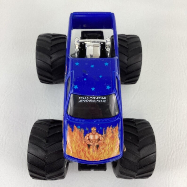 Op de afbeelding: Een blauw monstertruckspeelgoed met grote zwarte banden. De truck heeft een vlammenontwerp aan de voorkant en een grafische afbeelding van een gespierde figuur. De tekst "TEXAS OFF-ROAD PERFORMANCE" is zichtbaar op de voorruit. Blauwe sterren versieren de bovenkant.