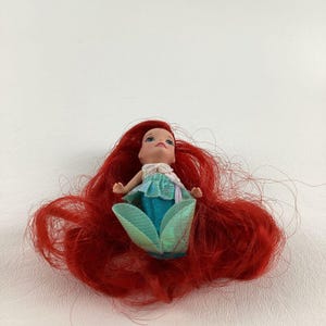Puede incluir: Una peque&ntilde;a mu&ntilde;eca de sirena con cabello rojo brillante, ojos azules y un atuendo turquesa y verde. La mu&ntilde;eca est&aacute; acostada, con su cabello extendido a su alrededor.