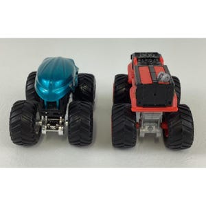 Pu&ograve; includere: Due monster truck giocattolo. Uno &egrave; verde acqua con cabina arrotondata, l'altro &egrave; rosso e nero con cabina piatta. Entrambi hanno grandi pneumatici neri e telaio argentato. I camion sono uno accanto all'altro.