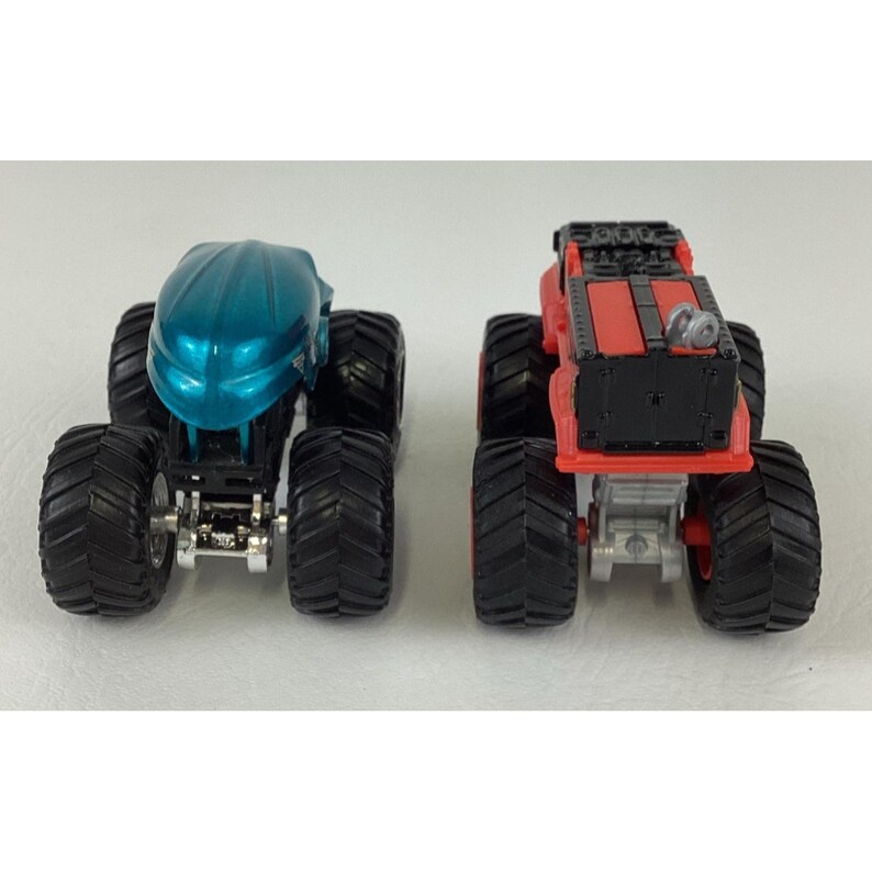 Pu&ograve; includere: Due monster truck giocattolo. Uno &egrave; verde acqua con cabina arrotondata, l'altro &egrave; rosso e nero con cabina piatta. Entrambi hanno grandi pneumatici neri e telaio argentato. I camion sono uno accanto all'altro.