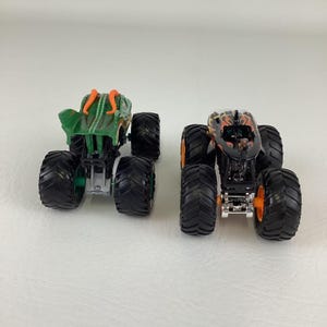 Puede incluir: Dos camiones monstruo de juguete. Uno es verde con detalles naranjas, el otro es negro con llamas naranjas. Ambos tienen neum&aacute;ticos negros grandes y est&aacute;n dise&ntilde;ados para el juego todoterreno.