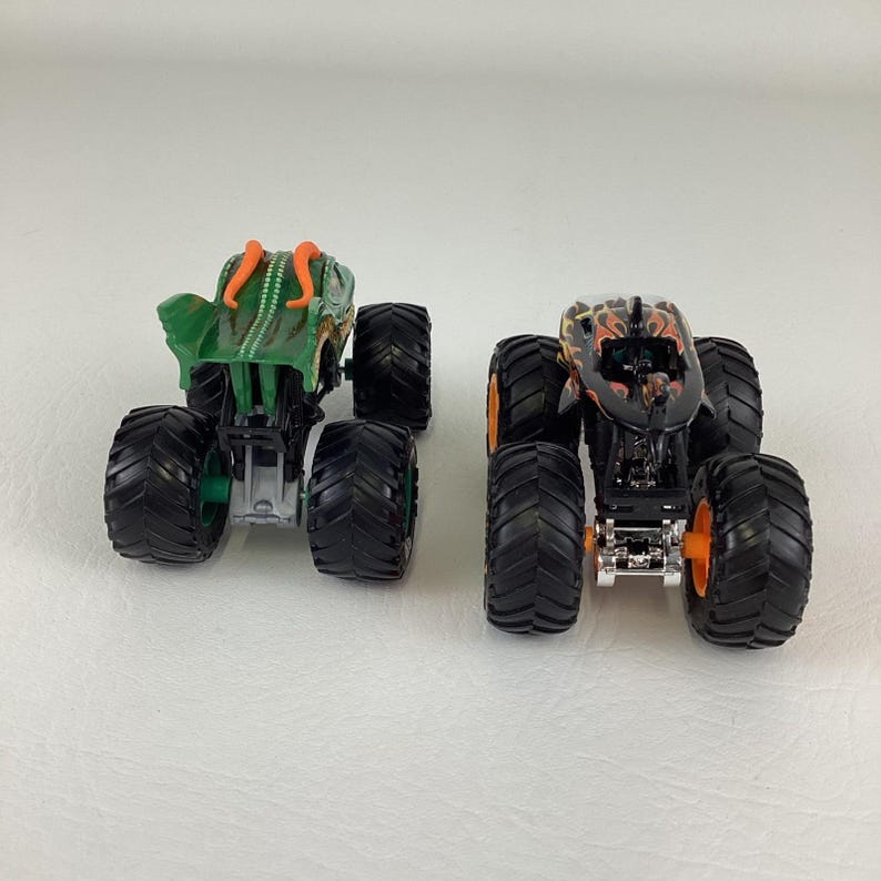 Puede incluir: Dos camiones monstruo de juguete. Uno es verde con detalles naranjas, el otro es negro con llamas naranjas. Ambos tienen neum&aacute;ticos negros grandes y est&aacute;n dise&ntilde;ados para el juego todoterreno.