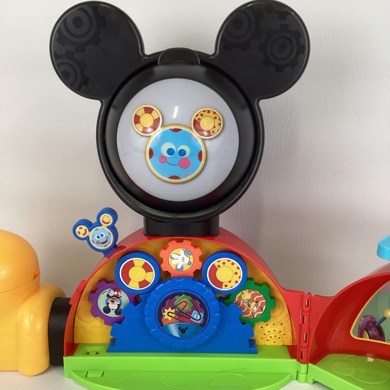 Pu&ograve; includere: Un colorato giocattolo di Mickey Mouse Clubhouse con una grande testa nera con dettagli a forma di ingranaggio. Il giocattolo ha un volto rotondo e illuminato con immagini di Mickey Mouse e vari ingranaggi ed elementi interattivi.
