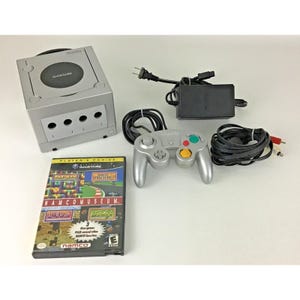 Puede incluir: Consola Nintendo GameCube plateada con un mando a juego, un adaptador de corriente y un cartucho de juego. El cartucho del juego está etiquetado como "Namco Museum" y presenta gráficos coloridos. El mando tiene botones verdes y amarillos.