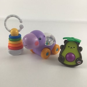 Pode incluir: Três brinquedos coloridos para bebés: um carro de hipopótamo roxo com uma janela de plástico transparente, um abacate verde com um caule castanho e um botão roxo, e um brinquedo de anéis empilháveis arco-íris com uma pega branca.