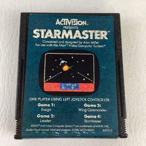 May include: A black Atari 2600 video game cartridge for the game Starmaster by Activision. The label features a dark teal background with white text, a space combat graphic, and lists game modes including Ensign, Leader, Wing Commander, and StarMaster.