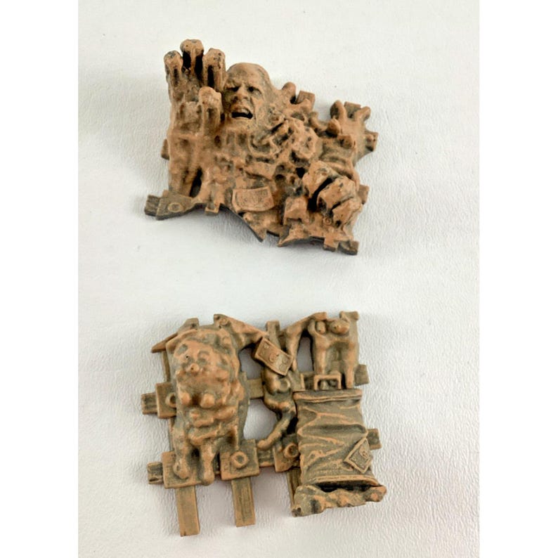 Puede incluir: Dos esculturas en miniatura marrones y texturizadas. La escultura superior representa una cara que emerge de una masa texturizada. La escultura inferior muestra una criatura con una valla y un objeto envuelto. Ambas est&aacute;n sobre un fondo blanco.