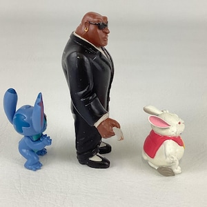 May include: Three character figurines: a blue alien, a man in a black suit, and a white rabbit in a red vest. The blue alien is in a crouching position. The man in the suit is wearing sunglasses. The rabbit has pink ears.