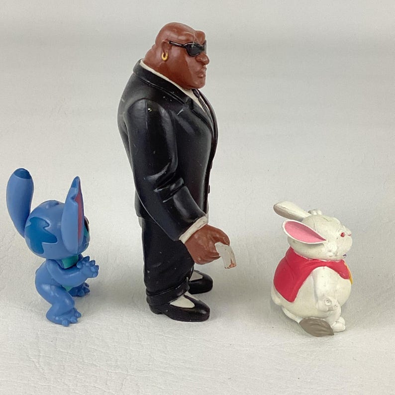 May include: Three character figurines: a blue alien, a man in a black suit, and a white rabbit in a red vest. The blue alien is in a crouching position. The man in the suit is wearing sunglasses. The rabbit has pink ears.