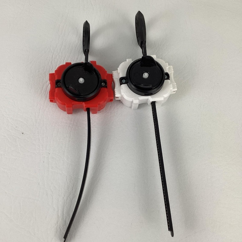 May include: Two toy launchers, one red and one white, each with a black center, pull cord, and handle. The launchers are designed for a spinning top game. The background is a plain white surface.