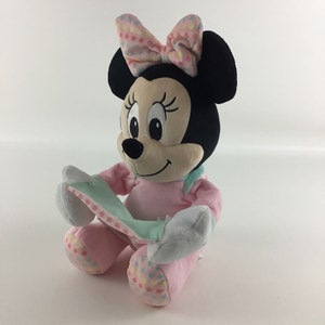 Op de afbeelding: Een pluche speelgoed van Minnie Mouse die een roze jurk draagt met kleurrijke stippen en een roze strik met kleurrijke stippen. Ze houdt een lichtblauwe en witte deken vast.