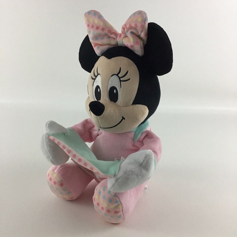 Op de afbeelding: Een pluche speelgoed van Minnie Mouse die een roze jurk draagt met kleurrijke stippen en een roze strik met kleurrijke stippen. Ze houdt een lichtblauwe en witte deken vast.