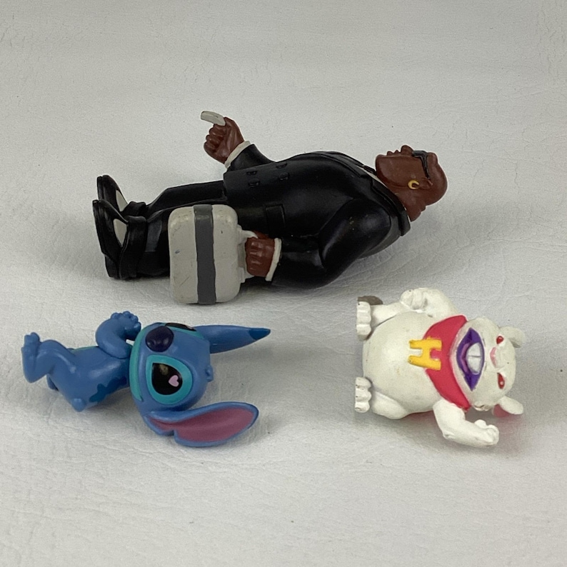 May include: Three small figurines: a figure in a black suit with a briefcase, a blue alien-like character, and a white rabbit-like figure with a red and yellow collar. The figures are lying on a white surface.