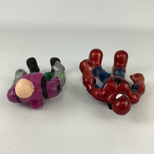 May include: Two superhero action figures. One is a red and blue Spider-Man figure. The other is a purple and green figure with a bald head. Both figures are lying on a white surface.