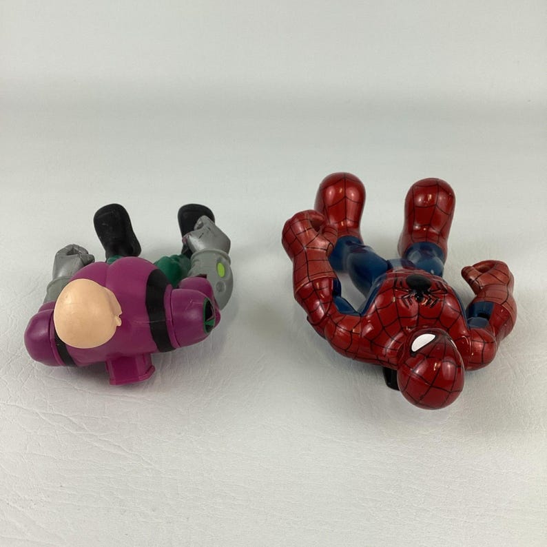 May include: Two superhero action figures. One is a red and blue Spider-Man figure. The other is a purple and green figure with a bald head. Both figures are lying on a white surface.