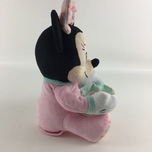 Op de afbeelding: Een pluche Minnie Mouse speelgoed dat een roze pyjama draagt en een groen-wit deken vasthoudt. Het speelgoed heeft een zwarte neus, grote zwarte ogen en een roze strik met witte stippen.