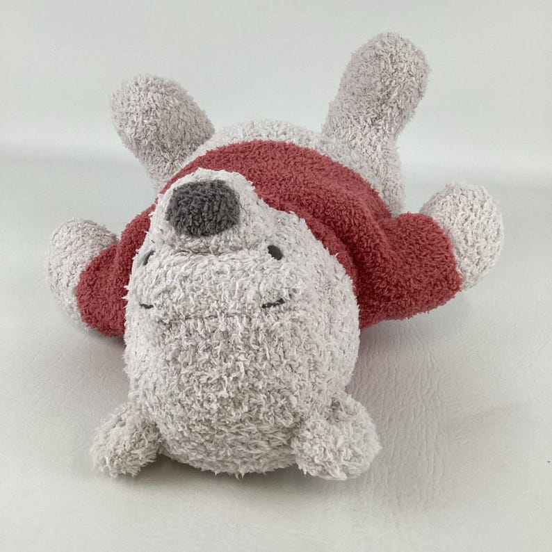 May include: A plush, light gray seal toy with a red sweater. The seal has a gray nose and closed eyes, lying on its back with its flippers and head up. The toy is soft and fluffy.