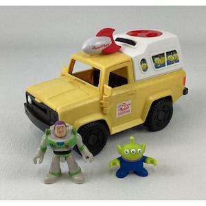 Könnte beinhalten: Ein gelber Spielzeug-Truck mit weißem Aufbau, einer rot-weißen Rakete auf dem Dach und Pizza Planet-Logos an der Seite. Der Truck hat schwarze Räder und Fenster mit grünen Alien-Grafiken. Zwei kleine Kunststofffiguren begleiten das Fahrzeug.